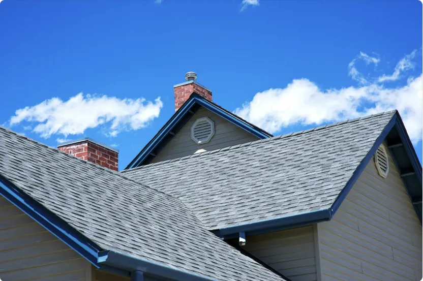 Close-up of a house roof with gray shingles, blue trim, and a brick chimney under a bright blue sky with white clouds.