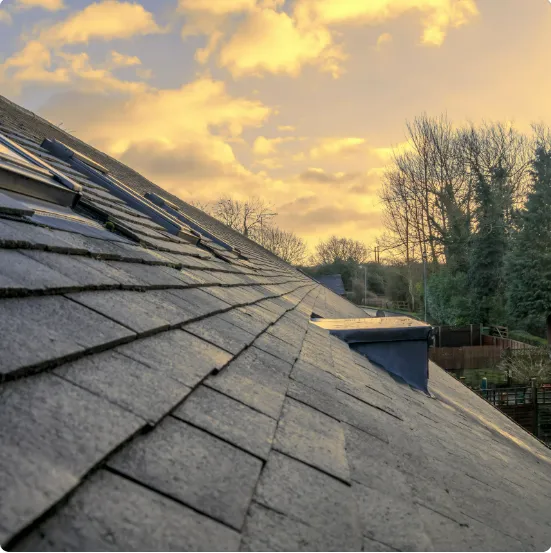 Sloped slate roof under a warm sunset sky with trees and nearby houses in the background.
