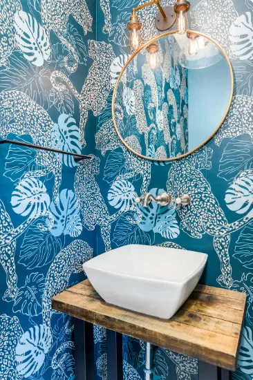 Bathroom with white square sink, wood plank counter, round mirror, and bold blue jungle wallpaper design.