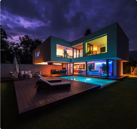 Modern night home with colorful interior lights, pool, wooden deck, and lounge area, perfect for entertaining.