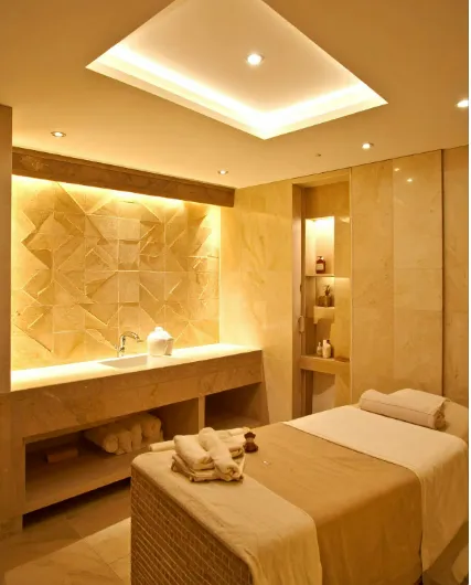 Serene spa room with warm lighting, massage table, folded towels, and stone sink, highlighting murals and decorative accents.