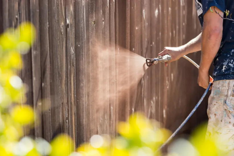 Person staining a wooden fence outdoors with spray paint, surrounded by flowers and greenery, ideal for arbor staining projects.