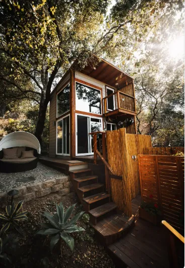 Modern wooden tiny house with balcony, large windows, and outdoor seating, surrounded by greenery for deck and arbor remodeling.