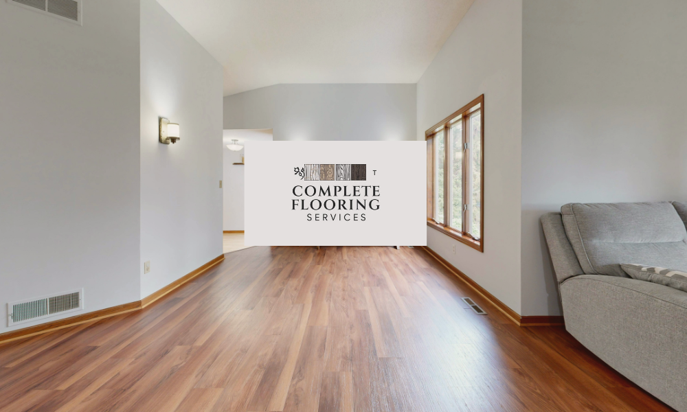 A bright, modern living room featuring professionally installed hardwood floors with a warm grain, with the 'Complete Flooring Services' logo overlaid in the center.