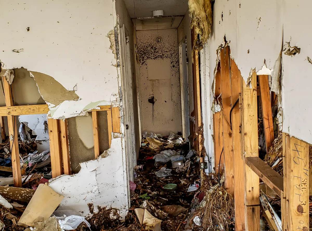 Flood-damaged hallway with torn drywall, exposed studs, and debris from structural water damage.