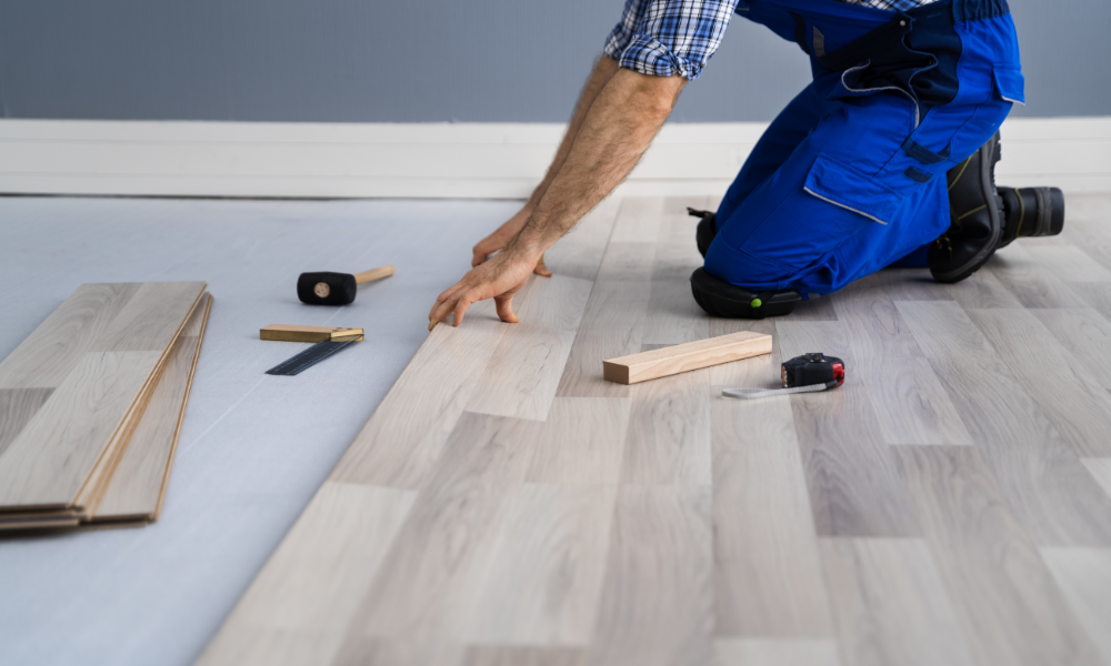 Our contractor in blue overalls is kneeling and installing wood laminate flooring with precision. Nearby, tools like a mallet and tape measure are scattered.