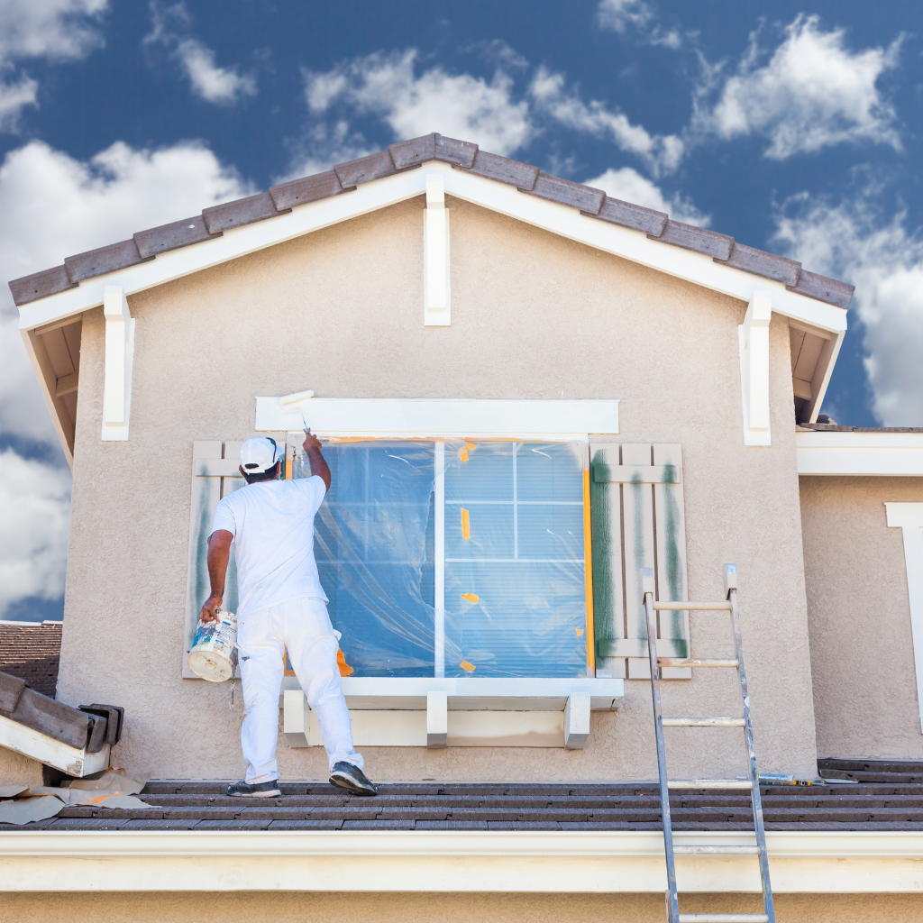 A professional painter in white uniform performing house painting on a residential exterior under a blue sky.
