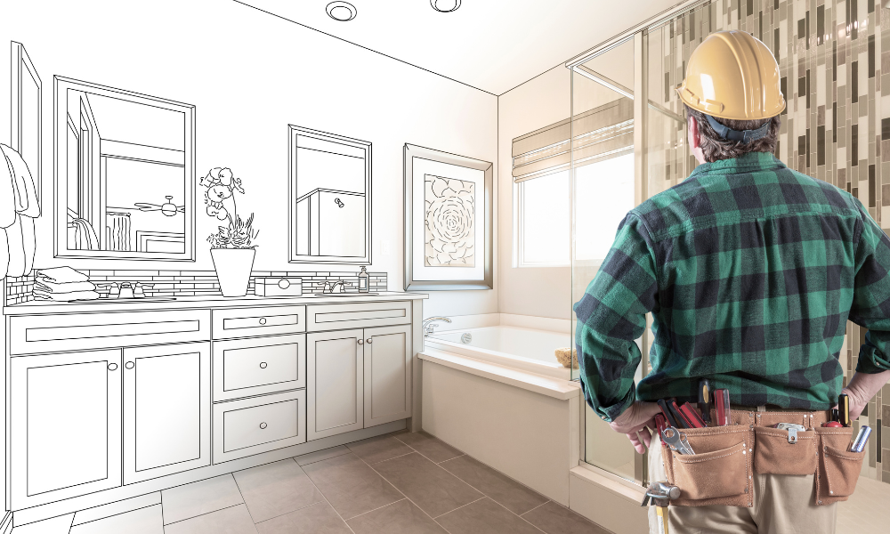 A licensed remodeling contractor with a tool belt and hard hat standing in a bathroom, looking at a digital wireframe sketch of a planned vanity and mirror renovation.