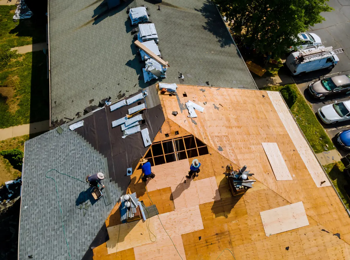Aerial view of a roof under construction. Workers lay shingles and wooden panels. Nearby parked cars and a tree indicate a residential area. Busy, sunny scene.