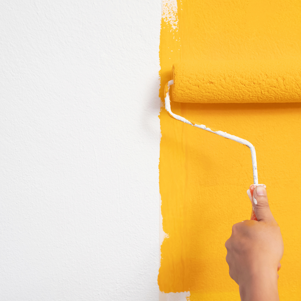 A hand using a paint roller to apply vibrant yellow paint over a white wall during a house painting project.