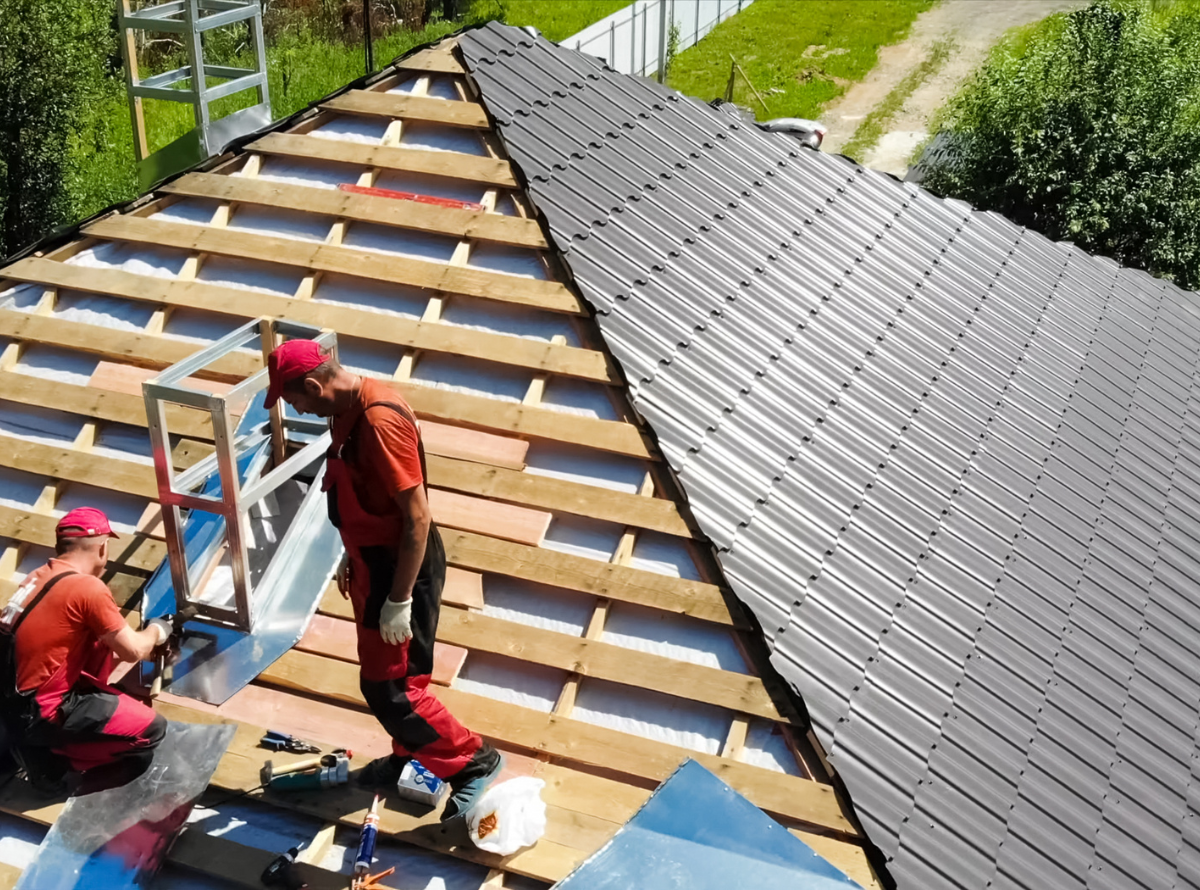 Two roofing contractors in red uniforms working on a residential roof replacement, installing new dark grey metal shingles over a wooden frame.