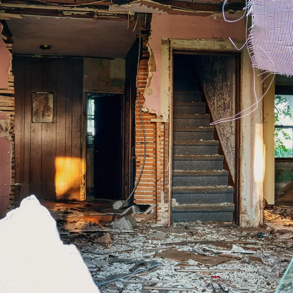 Dilapidated room with torn wallpaper, exposed wood, and debris on the floor. Stairs lead up on the right; sunlight casts a warm glow, highlighting decay.